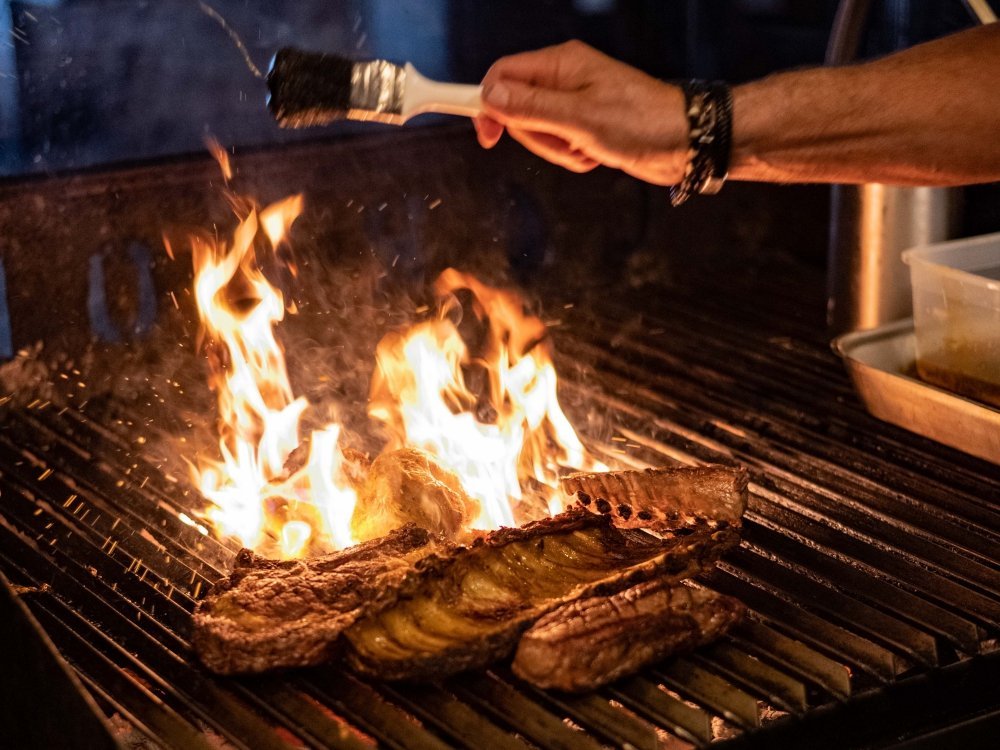 Steak en ribs op het open vuur bij Cafe Carbon, vlammen slaan op uit de grill in een donkere keuken
